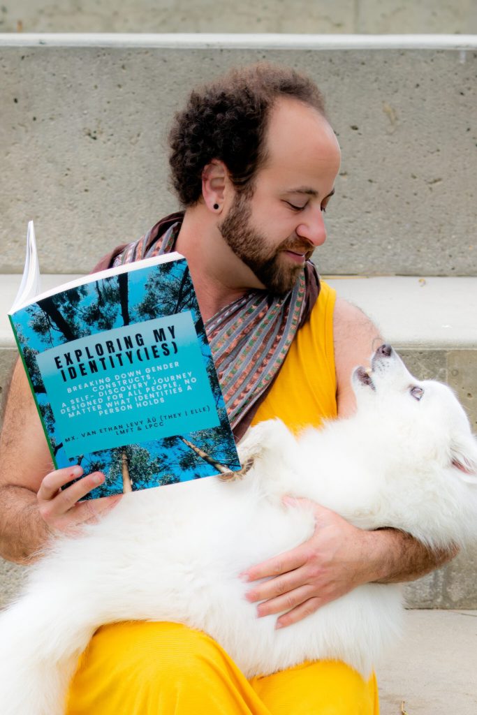 This image contains an olive skin person who has a brown beard, curly brown hair while looking down at their a white dog on their lap, wearing a yellow dress with a scarf around their neck, and smiling at their dog. In their hand is a copy of their interactive workbook, Exploring My Identity(ies) in bold black letters. The next line shows breaking down gender constructs, a self-discovery journey designed for all people, no matter what identities a person holds in bold white letters. The next line shows Mx. Van Ethan Levy, AÜ (They | Elle) LMFT & LPCC in bold black letters. All the words are in a blue box. The blue box is surrounded by tall green trees all creating a circular illusion. Van and Sushi are sitting on some grey steps.