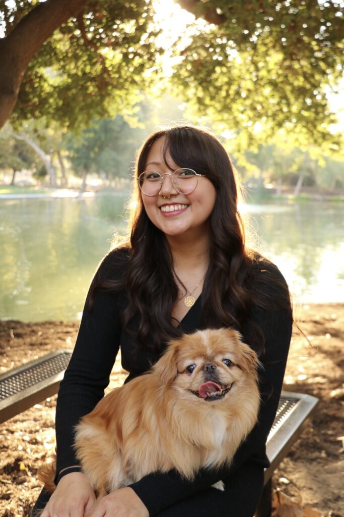 A medium skinned fem presenting person with long black hair, wearing glasses. They use she pronouns. She is wearing a long sleeved black dress. She has a gold horseshoe septum piercing. She is wearing round thin gold frame glasses. She is sitting outside on a bench in front of a lake with trees above. She is also holding a small golden long-haired dog with its tongue sticking out.